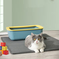 Cat lying on a mat next to a blue litter box with yellow interior in a room.